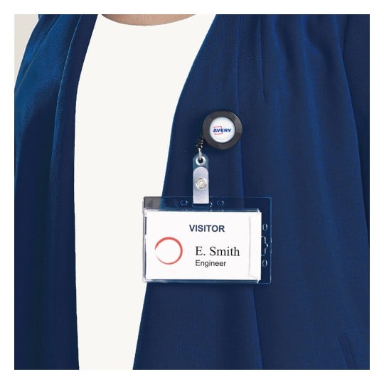 Das Bild zeigt einen Besucher-Namensschild, das an der Kleidung einer Person befestigt ist. Das Schild trägt den Namen 'E. Smith' und den Titel 'Ingenieur.' Es zeigt außerdem einen Anstecker mit dem Avery-Logo.