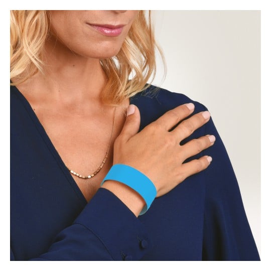 Das Bild zeigt eine Frau mit blonden Haaren, die eine dunkelblaue Bluse trägt. Sie berührt ihr Handgelenk, an dem sich ein leuchtend blaues Armband befindet.
