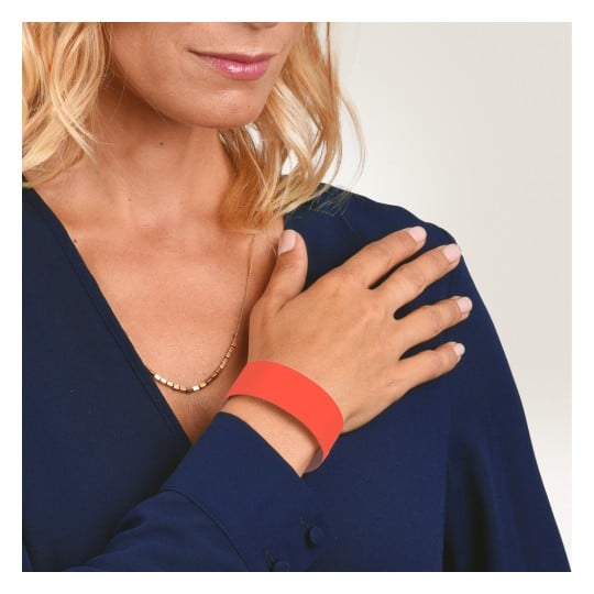 Das Bild zeigt eine Frau mit blonden Haaren, die eine dunkelblaue Bluse trägt und eine Hand auf ihrer Schulter platziert hat. Sie hat eine dünne Halskette und trägt ein orangefarbenes Armband am Handgelenk.