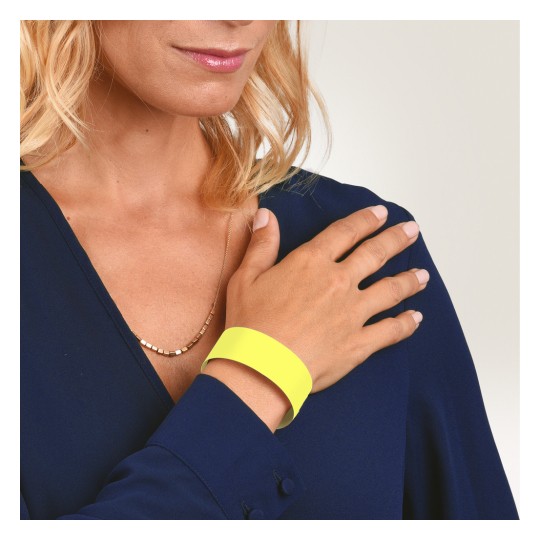Das Bild zeigt eine Frau mit blondem Haar, die ein dunkelblaues Hemd trägt. Sie ist mit einer goldenen Halskette geschmückt und trägt ein leuchtend gelbes Armband am Handgelenk, wobei ihre Hand sanft auf ihrer Schulter ruht.
