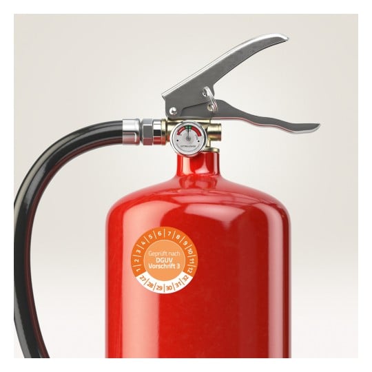 Das Bild zeigt einen roten Feuerlöscher mit einer Metalldüse, einem Druckmesser und einem Etikett, das die Einhaltung der Sicherheitsstandards anzeigt.