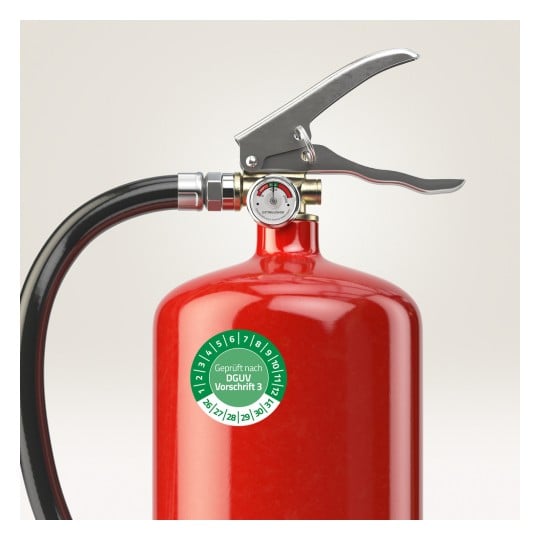 Das Bild zeigt einen roten Feuerlöscher mit einem schwarzen Schlauch und einem Manometer. Es gibt ein Etikett, das die Einhaltung der Sicherheitsvorschriften angibt.