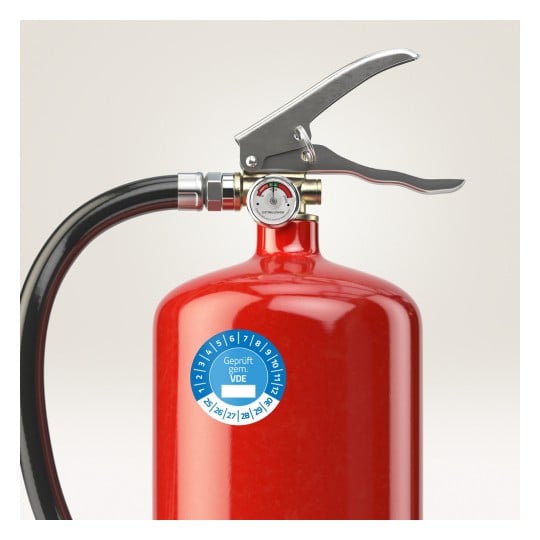 Das Bild zeigt einen roten Feuerlöscher mit einem Manometer und einer Düse. Das Manometer zeigt den Druckpegel an, und es gibt einen blauen Aufkleber, der die Zertifizierung angibt.