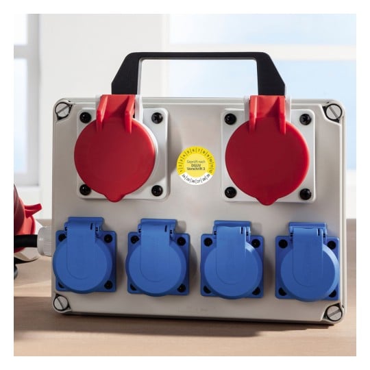 Das Bild zeigt eine Stromverteilungsbox mit zwei roten runden Steckdosen oben und vier blauen runden Steckdosen unten. Sie ist mit einem Griff für den Transport ausgestattet und besitzt einen gelben Zertifizierungsaufkleber, der anzeigt, dass sie den Sicherheitsstandards entspricht.