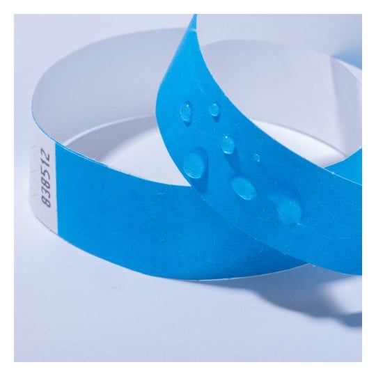 Das Bild zeigt zwei blaue Identifikationsarmbänder, wobei eines das andere überlappt, mit Wassertropfen auf ihrer Oberfläche. Sie scheinen aus einem papierähnlichen Material zu bestehen und haben auf einer Seite einen Aufkleber mit Nummern.
