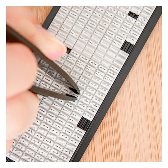 Das Bild zeigt eine Hand, die mit einer Pinzette Buchstaben aus einem Satz kleiner weißer Plastikplättchen entnimmt, die in Reihen angeordnet sind, mit einer hölzernen Oberfläche darunter.