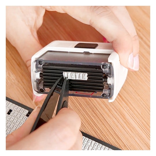 Das Bild zeigt eine Person, die mit einer Pinzette ein kleines Objekt in einem Handgerät oder Stempel handhabt. Im Hintergrund ist eine Holzoberfläche zu sehen, auf der weitere Stempel oder Buchstaben sichtbar sind.
