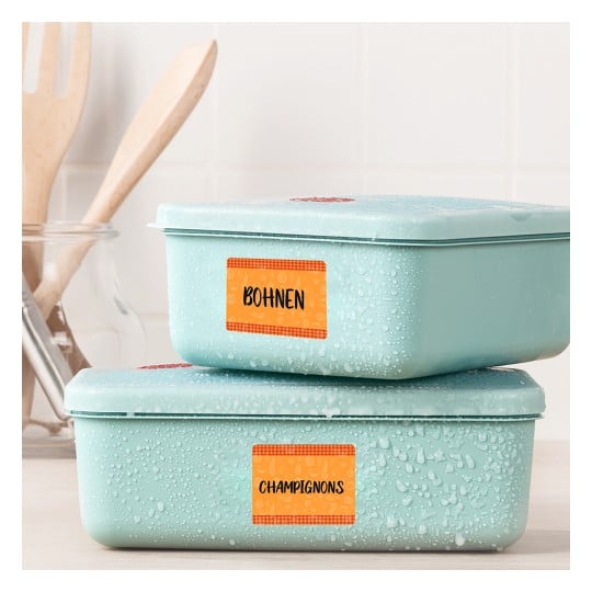 Das Bild zeigt zwei übereinander gestapelte, hellblaue Aufbewahrungsbehälter mit Etiketten. Der obere Behälter ist mit 'Bohnen' und der untere mit 'Champignons' beschriftet. Auf den Behältern sind Wassertropfen sichtbar, und im Hintergrund sind Holzbestecke unscharf zu erkennen.