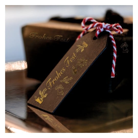 Das Bild zeigt ein hölzernes Geschenkanhänger mit dem goldenen Text 'Frohes Fest'. Der Anhänger ist mit festlichen Symbolen wie einer Schneeflocke, Glocken, einer Kerze und immergrünen Zweigen verziert und mit rotem und weißem Garn gebunden.