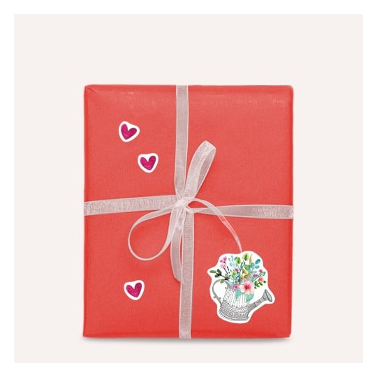 Das Bild zeigt ein schön verpacktes Geschenk in korallfarbenem Papier mit einem silbernen Band. Es ist mit dekorativen Aufklebern von Herzen und einer Blumen-Gießkanne versehen.