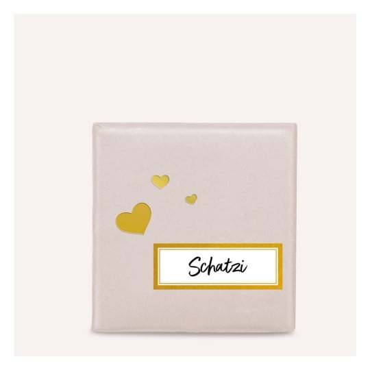 Das Bild zeigt eine quadratische Geschenkbox, die in einem sanft gefärbten Papier eingewickelt ist. Sie hat ein Etikett mit dem Wort 'Schatzi' in einer stilvollen Schriftart und kleinen goldenen Herzen, die darum schweben.