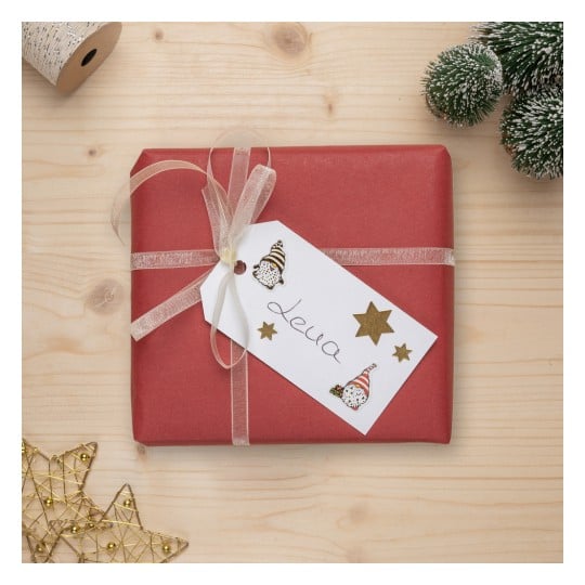 Das Bild zeigt eine wunderschön verpackte rote Geschenkschachtel mit einem transparenten Band und einem Geschenkanhänger, der einen handgeschriebenen Namen enthält. Auf dem Anhänger befinden sich dekorative Elemente wie Sterne. In der Nähe gibt es eine kleine immergrüne Dekoration und einen sternförmigen Anhänger, die alle auf einer hölzernen Oberfläche platziert sind.