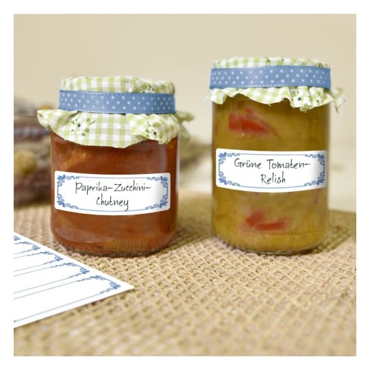 Das Bild zeigt zwei Gläser mit hausgemachten Würzmitteln. Ein Glas enthält 'Paprika-Zucchini-Chutney', während das andere 'Grüne Tomaten-Relish' enthält. Beide Gläser sind mit dekorativen Deckeln versehen und stehen auf einer Sackleinen-Oberfläche.