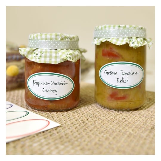 Das Bild zeigt zwei Gläser mit Eingemachtem. Ein Glas enthält Paprika-Zucchini-Chutney und das andere enthält grünes Tomaten-Relish. Beide Gläser sind mit karierten Stoffdeckeln versehen.