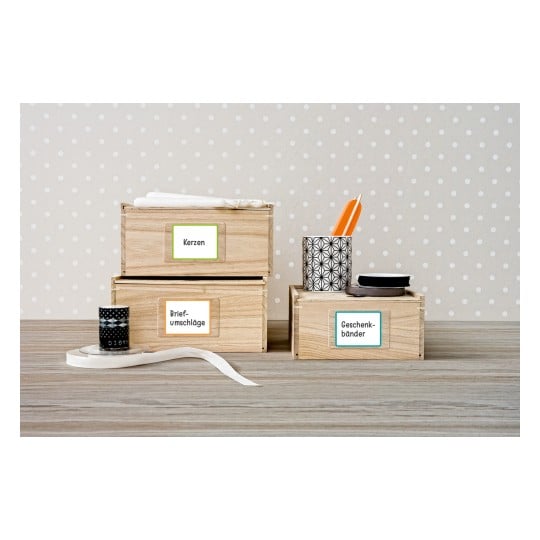 Das Bild zeigt drei Holzkisten mit deutschen Beschriftungen: 'Kerzen', 'Briefumschläge' und 'Geschenkebänder'. In der Nähe befinden sich auch dekorative Gegenstände wie Klebeband und Stifte.