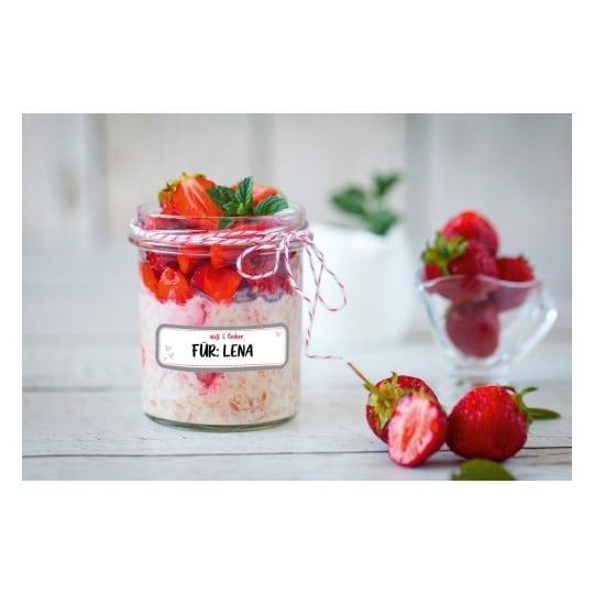 Das Bild zeigt ein Glas, das mit einem geschichteten Dessert aus Haferflocken und Erdbeeren gefüllt ist, gekrönt mit frischen Erdbeerstücken und einem Minzblatt. Das Glas hat ein Etikett mit der Aufschrift 'FÜR: LENA'. Neben dem Glas liegen einige ganze Erdbeeren.