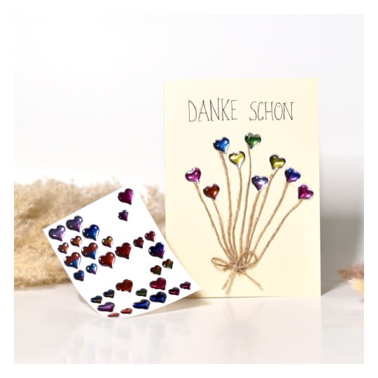 Das Bild zeigt eine Grußkarte mit dem Text 'Danke Schön' und farbige Herzformen, die an Schnüren befestigt sind. Es gibt auch ein Stickerblatt mit verschiedenen Herzstickern.
