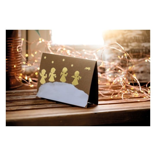 Das Bild zeigt eine Weihnachtskarte mit goldenen Silhouetten von Engeln auf einem braunen Hintergrund, die auf einer Holzoberfläche platziert ist, mit sanft leuchtenden Lichtern im Hintergrund.