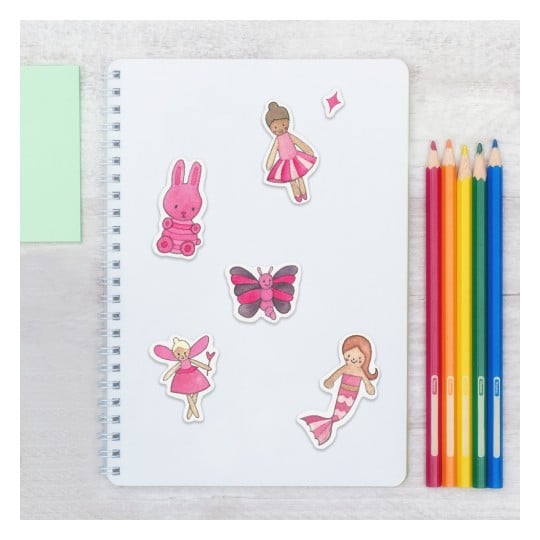 Das Bild zeigt ein weißes Spiralblock, geschmückt mit bunten Aufklebern, darunter ein rosa Hase, verschiedene Puppen, ein Schmetterling und eine Meerjungfrau. Rechts sind mehrere Buntstifte in einer Reihe angeordnet.