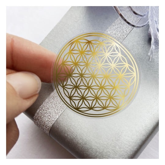 Das Bild zeigt eine Nahaufnahme einer Hand, die einen goldenen Aufkleber mit Blumenmuster auf eine silberne Geschenkbox drückt, die mit einem glitzernden Band verziert ist.