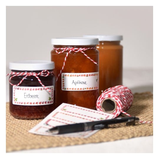 Das Bild zeigt drei Gläser Marmelade mit den Etiketten 'Erdbeere' und 'Aprikose', dekorativem Bindfaden und Etiketten in der Nähe, auf einem Jutetuch arrangiert.