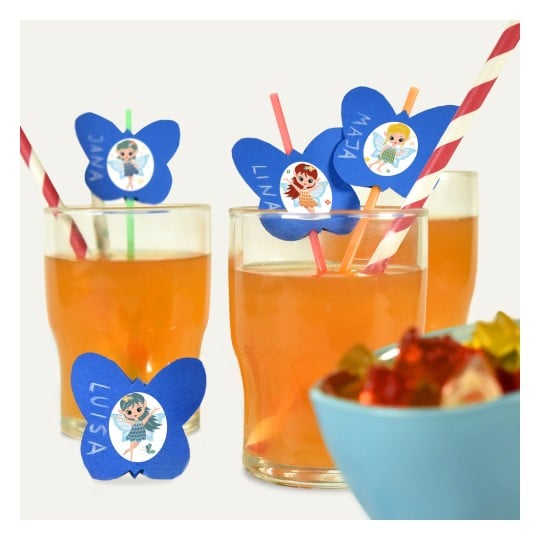 Das Bild zeigt mehrere Gläser mit orangefarbenen Getränken, die mit bunten, schmetterlingsförmigen Dekorationen und Strohhalmen verziert sind. Jede Dekoration hat einen Namen und eine Illustration einer Figur. Im Vordergrund befindet sich auch eine Schüssel mit bunten Gummibärchen.