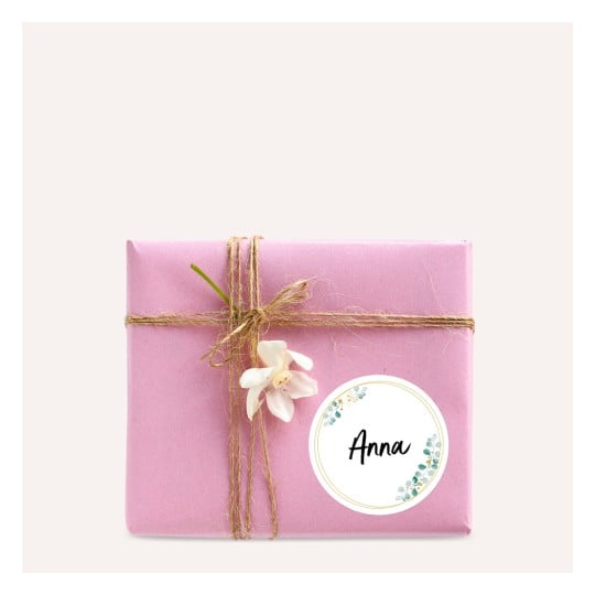 Das Bild zeigt ein wunderschön verpacktes rosa Geschenkbox, verziert mit Schnur und einer kleinen Blume. Ein runder Anhänger mit dem Namen 'Anna' ist an das Paket angebracht.