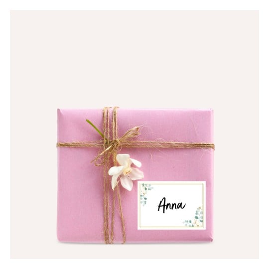 Das Bild zeigt ein rosa eingepacktes Geschenk mit Kordel darum und einer angehefteten Blume. Es gibt ein Etikett, auf dem 'Anna' steht.