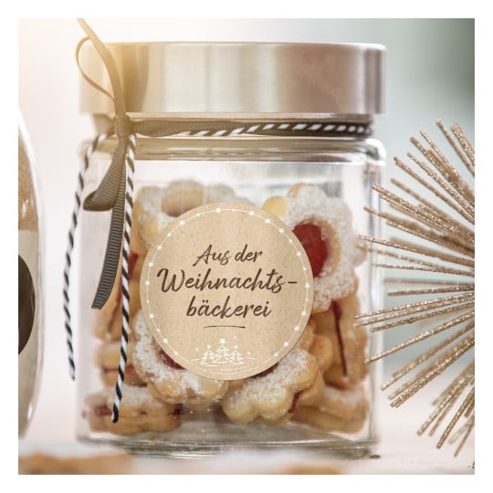 Das Bild zeigt ein Glasgefäß, gefüllt mit festlichen Keksen, beschriftet auf Deutsch mit 'Aus der Weihnachtsbäckerei', was darauf hinweist, dass sie aus der Weihnachtsbäckerei stammen. Das Gefäß ist elegant mit einem Band verziert, und in der Nähe befinden sich dekorative Elemente, einschließlich eines sternförmigen Ornaments.