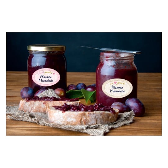 Das Bild zeigt zwei Gläser Pflaumenmarmelade, Brotscheiben, die mit der Marmelade bestrichen sind, und ein paar Pflaumen auf einer Holzoberfläche. Die Gläser haben Etiketten auf Deutsch, und die Szene ist vor einem dunklen Hintergrund arrangiert.