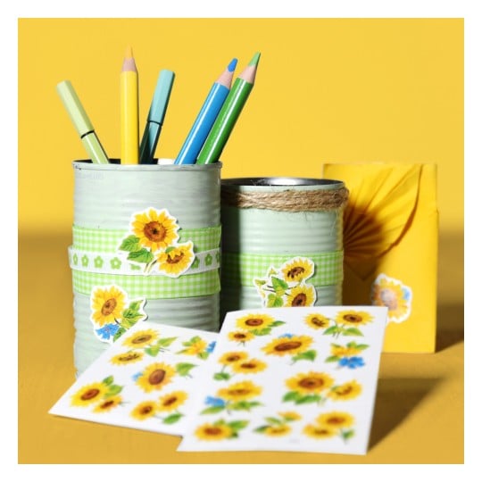 Das Bild zeigt zwei verzierte Blechdosen, die Buntstifte halten. Die Dosen sind mit Sonnenblumenaufklebern und grünen karierten Bändern verziert und vor einem leuchtend gelben Hintergrund platziert. Neben den Dosen liegen Bögen mit Sonnenblumenaufklebern, die der Szene eine fröhliche und künstlerische Atmosphäre verleihen.