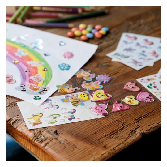 Das Bild zeigt einen kreativen Arbeitsbereich mit bunten Zeichnungen und verschiedenen Aufklebern. Ein Regenbogen ist auf Papier gemalt und mit glänzenden Edelsteinen dekoriert. Es gibt mehrere Bögen mit Aufklebern von Cartoon-Charakteren, Herzen und Blumen, zusammen mit Buntstiften und verstreuten Süßigkeiten auf einem Holztisch.