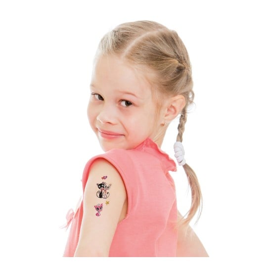 Ein junges Mädchen mit einem spielerischen Ausdruck schaut zurück und zeigt temporäre Tattoos auf ihrem Arm. Sie hat hellblondes Haar, das zu Zöpfen geflochten ist, und trägt ein rosa Shirt.