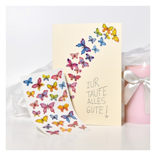 Das Bild zeigt eine Grußkarte und ein Blatt mit Schmetterlingsstickern. Die Karte in hellem Cremefarben hat ein dekoratives Design mit bunten Schmetterlingen entlang der Oberseite und zeigt auch den deutschen Satz 'Zur Taufe alles Gute!'. Eine rosa Kerze mit einer Schleife ist neben der Karte platziert.