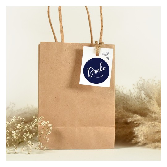 Das Bild zeigt eine braune Papiertüte mit einem Anhänger, auf dem 'Danke' steht. Neben der Tasche liegen getrocknete Blumen, die zu einer rustikalen Ästhetik beitragen.