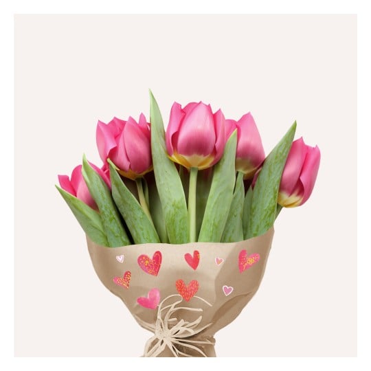 Ein Strauß rosa Tulpen in Papier mit dekorativen Herzen gewickelt, vor einem hellen Hintergrund platziert.