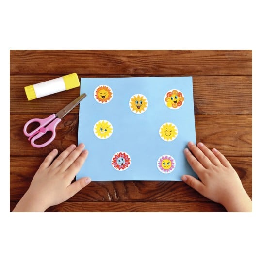 Das Bild zeigt ein blaues Papier mit bunten Sonnenstickern darauf, eine rosafarbene Schere und einen Klebestift, die auf einem Holztisch liegen. Die Hände eines Kindes sind sichtbar, eine Hand berührt das Papier mit den Stickern, während die andere die Schere hält.