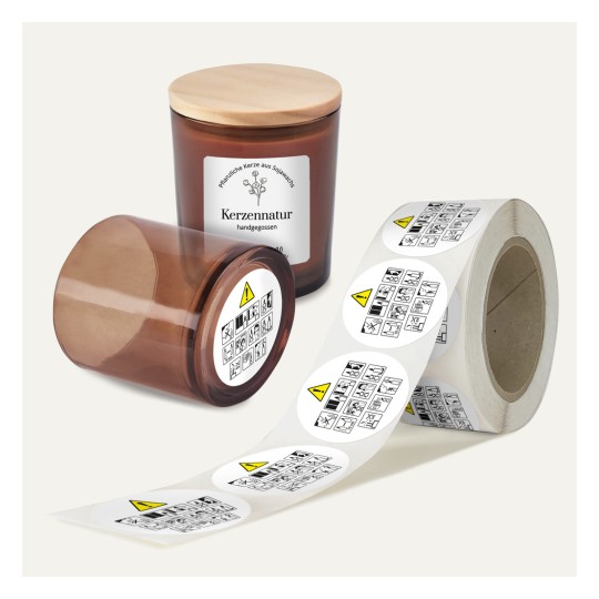 Das Bild zeigt zwei braune Glaskerzengefäße mit Holzdeckeln, die mit 'Kerzennatur' beschriftet sind, sowie eine Rolle mit Warnetiketten, die verschiedene Symbole aufweisen. Ein Gefäß ist geneigt und das andere steht aufrecht.