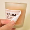 Von einer Kerze wird ein bedrucktes Kerzenetikett abgelöst. Das Material ist die ablösbare, weiße Folie.
