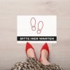 Auf dem Boden klebt ein Bodenaufkleber auf dem zwei Schuhabdrücke zu sehen sind und "Bitte hier warten" steht. Davor steht eine Frau.