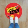 Ein runder, roter Bodenaufkleber klebt am Boden und darauf steht "Begrenzte Zeit Super Sale". Jemand steht mit weißen Sneakers halb auf dem Bodenaufkleber.