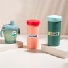 Three colorful beverage containers arranged on a surface. The left container is a sippy cup, the middle is a peach-colored drink container, and the right is a dark green tumbler, all labeled with 'ENZO BOYER'.