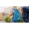 Ein junges Kind mit einem blauen Rucksack mit Raketenmotiv hält ein Buch in der einen Hand und die Hand eines Erwachsenen in der anderen, während es auf einem Weg in der Nähe von Gebäuden und Grünflächen geht.