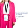Das Bild zeigt eine Person, die eine leuchtend pinke Anzugjacke und Hose trägt und ein Abzeichen hält, auf dem ihr Name, ihre Rolle und Details zu einer Veranstaltung stehen. Es gibt eine Nachricht, die darauf hinweist, dass das Abzeichen für mehrere Veranstaltungen wiederverwendbar ist.