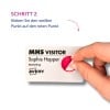 Das Bild zeigt eine Hand, die einen weißen Aufkleber auf einem Besucher-Ausweis anbringt, der den Namen Sophie Hopper und den Bereich Marketing enthält. Es enthält Anweisungen auf Deutsch, einen weißen Punkt auf einen roten Punkt zu setzen.