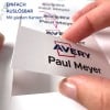 Das Bild zeigt eine Hand, die ein Etikett mit dem Namen 'Paul Meyer' neben dem 'Avery'-Logo hält. Es gibt einen Text, der darauf hinweist, dass das Etikett leicht ablösbar ist und glatte Kanten hat.