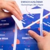 Das Bild zeigt Hände, die eine bedruckte Kartonkarte mit Namen und Bezeichnungen halten, wobei ein Merkmal der einfachen Entfernung hervorgehoben wird und ein farbenfrohes Design zu sehen ist. Der Text auf Deutsch erwähnt hochwertiges Papier.