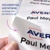 Das Bild zeigt eine Nahaufnahme eines Satinetiketts mit dem Text 'Avery Paul Meyer' und einer Beschreibung auf Deutsch über das Material, die darauf hindeutet, dass es gut auf Kleidung passt.