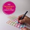 Das Bild zeigt eine Hand, die einen Permanentmarker benutzt, um bunte Haftnotizen oder Etiketten mit dem Text 'TINA S.' zu beschriften. Der Hintergrund ist neutral, und der Text auf dem Bild ist auf Deutsch, was darauf hinweist, dass diese Etiketten mit einem Permanentmarker beschriftet werden sollen.