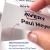 Das Bild zeigt eine Nahaufnahme einer Hand, die ein weißes Etikett mit dem Namen „Paul Meyer“ und dem prominent dargestellten „Avery“-Logo hält. Das Etikett scheint Teil einer Produktbeschreibung auf Deutsch zu sein, in der von glatten Kanten und hochwertigem Papier die Rede ist.